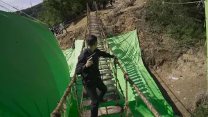 Magic Media VFX, A man holding a pistol crosses a rope bridge on a film set with green screens below. Crew members are in the background.